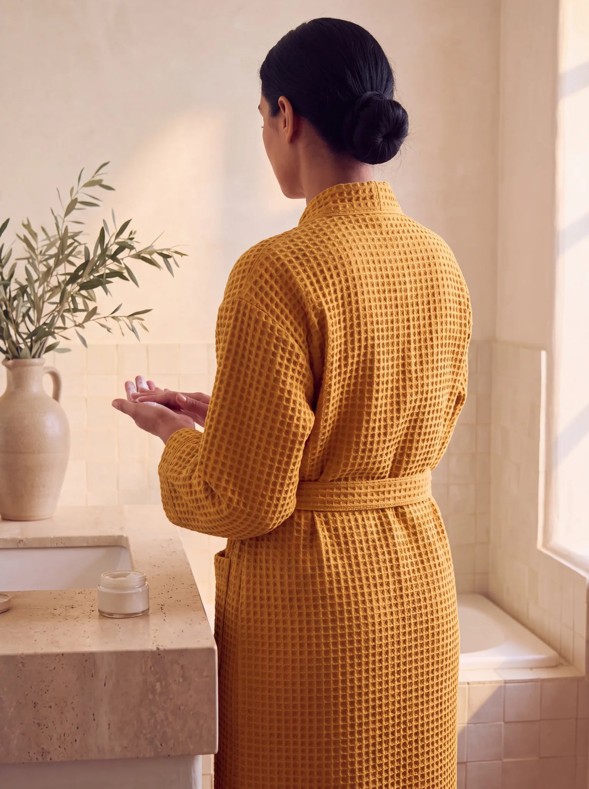 Femme de dos portant le peignoir Lumä en nid d’abeille jaune devant un lavabo